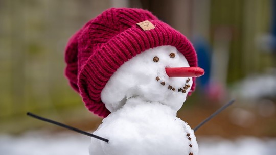 A close-up of a snowman's head. He is wearing a red wool hat, has a red crayon for a nose and a big smile