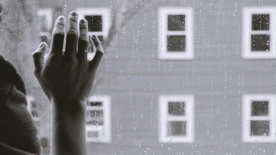 A black and white image of a woman's hand against a window, outside the window is spattered with rain