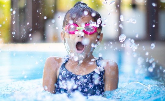 A young girl in pink swimming goggles emerging from the water in a pool with a splash