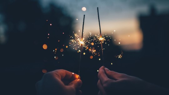 Two hands holding sparklers, the background is blurred, but the sun has just set and the sky is dark blues and orange