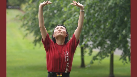 A woman stretching her arms above her head. She is outdoors in a green space with trees.