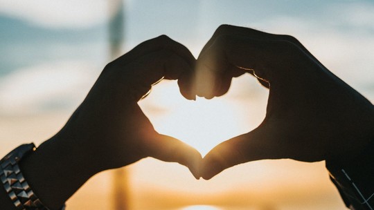A close up of two hands making a heart shape with the sun setting in the background