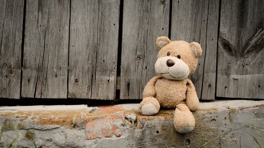 A well-loved toy bear sitting on a step next to a wooden fence. The bear looks abandoned.