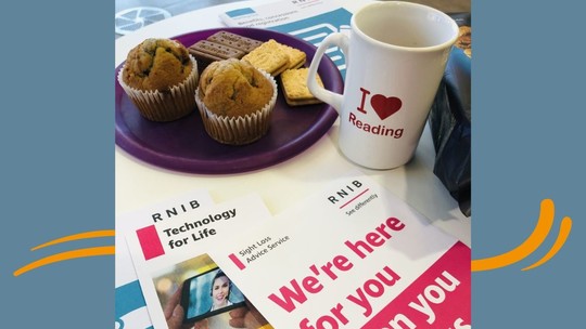 A mug and a plate of small cakes on a table along with some visual impairment information leaflets