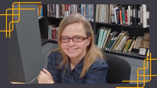 A head and shoulders photo of Charlotte Dade, smiling, sitting at a desk with bookshelves in the background