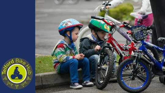 2 small boys in bike helmets sitting on the kerb. Their bikes have training wheels and are stood to the right of the picture.