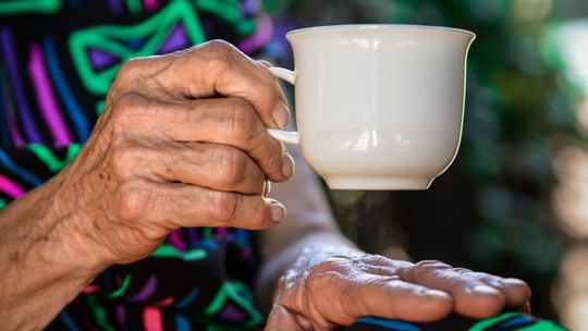 A close-up of an older person's hand holding a white porcelain cup of tea