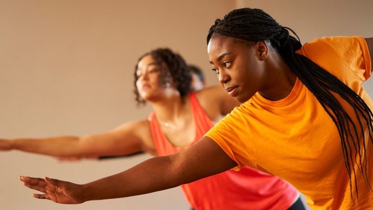 Two women in bright t-shirts do a standing stretch with their right arms forward