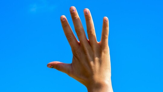 A hand, held up to a clear blue sky. The nails of the hand have small rainbows painted on each of them.