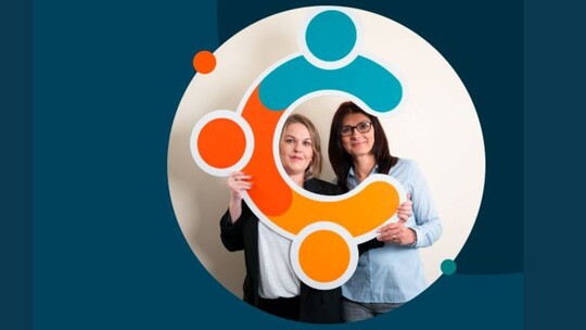 2 women holding up a big cardboard C, which is the Cancer Champions logo, so that they are framed by it.  