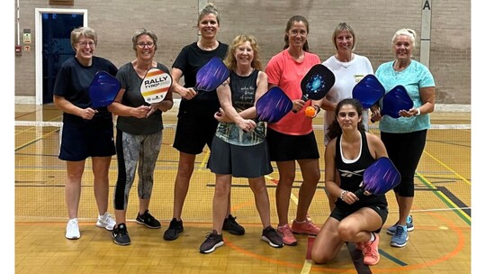 A group of 8 smiling women or various ages in sports clothes, posing with the Pickleball paddles.