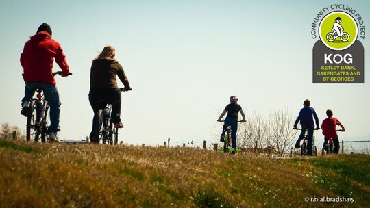 2 adults and 3 young people on bikes cycling away from the camera along a green track. The KOG Cycling Project logo is in the top right corner.