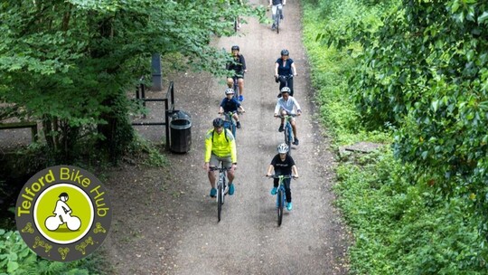A group of 7 people cycling through a woodland area. The Bike Hub logo appears in the bottom left corner.