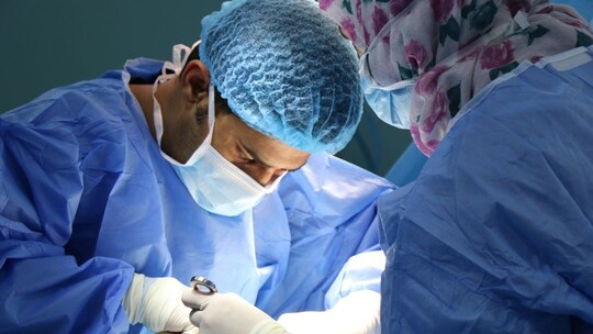 Two doctors leaning over an operating table, wearing blue scrubs, caps, masks and rubber gloves.