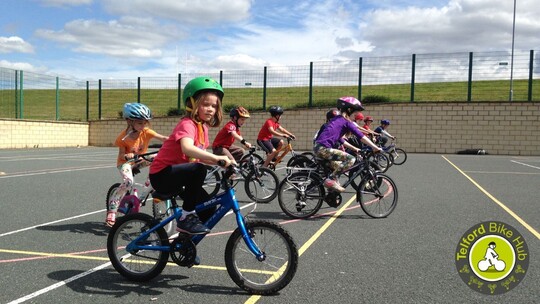 9 primary age children in cycle helmets riding their bikes across a sports court at Hadley Learning Community 