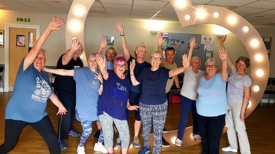 An enthusiastic, laughing group of 11 older people in t-shirts and leggings, all with their hands in the air