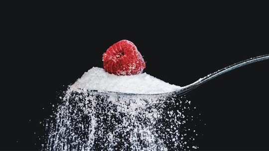 A spoon heaped with sugar and a raspberry perched on top. The background of the image is black.