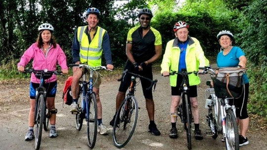A group of 5 cyclists of varying ages standing astride their bikes in a wooded lane