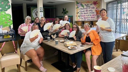 A group of 10 ladies sitting around a cafe table. They are all posing with paper fans open in front of them.