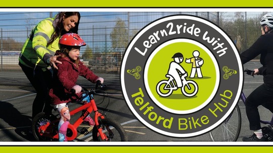 A woman in a high-vis vest helps a small girl in bike helmet, as she rides her bike. The Telford Bike Hub logo is in the right of the picture.