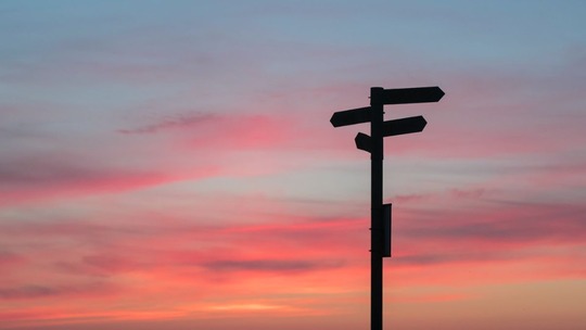 The silhouette of a four-way signpost against a sunset sky of orange, pink and blue