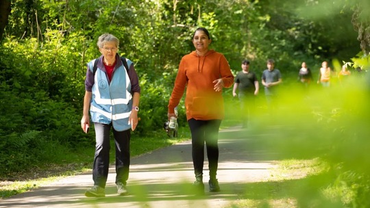 2 women, one in a walk leader high-vis vest, walking on a sunny path surrounded by trees