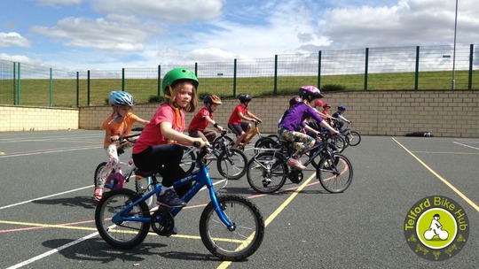 9 children wearing helmets and on bikes, riding across an outdoor sports court area on a sunny day.
