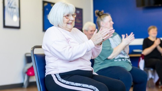 A group of older people doing seated exercises in a community room. All are smiling, while doing an arm exercise.