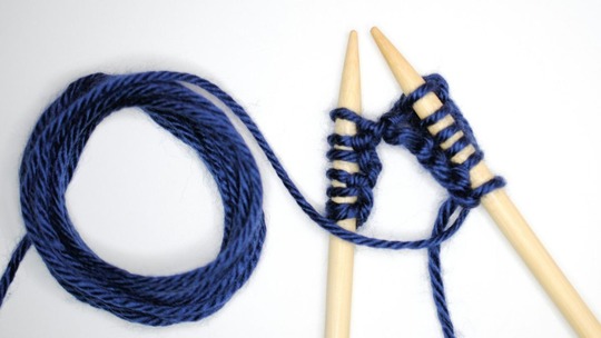 A skein of blue wool and the beginnings of some knitting on two large knitting needles.