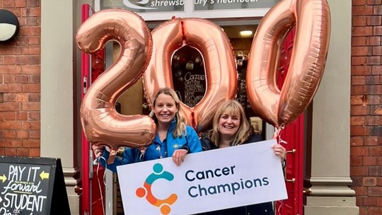 2 women, holding gold balloons in the shape of 2, 0 and 0 (200) and a large Cancer Champions logo.