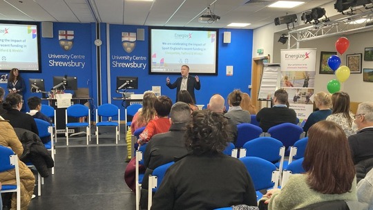 Photo of a man speaking at the front of a room of seated people.