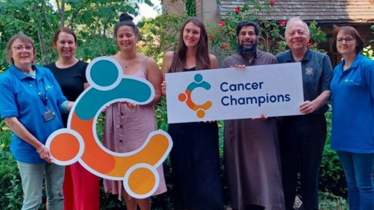 A group of smiling men and women of all ages hold up the Cancer Champions logo and sign