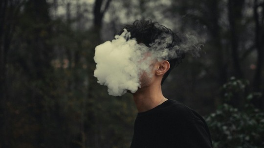 Head and shoulders image of a young man in dark woodland area. His head is completely engulfed in a cloud of smoke.