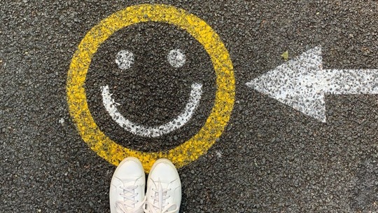 A yellow smiley face painted on some pavement with a white arrow pointing toward it. Two feet stand at the end of the image.