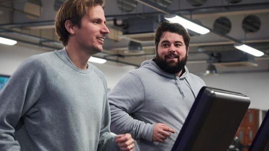 2 men in their 20s side by side on running machines in a gym. Both wear grey sweatshirts.