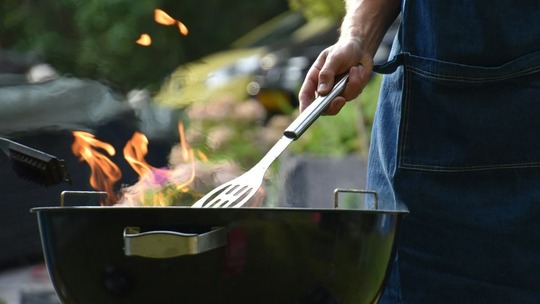 Close up photo of a barbecue and a man's torso. The man holds some tongs and is wearing an apron.