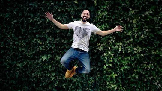 A happy man of about 30 years old jumps up, his arms out wide, against a backdrop of greenery