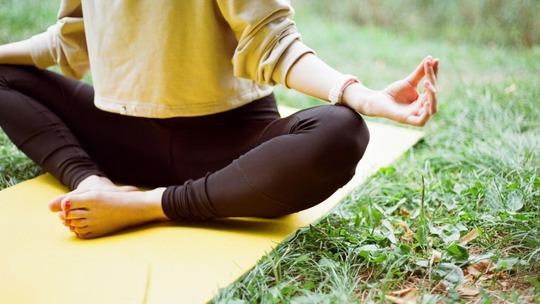 A person sitting cross legged on a yoga mat on the grass. We can only see then from their torso downwards.