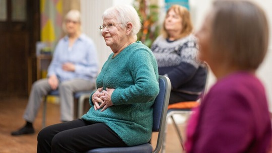 An older woman doing chair exercises in a room with a number of other older people