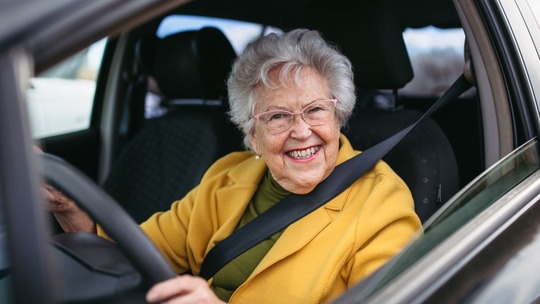 An older woman in a yellow coat, sitting at the wheel of her car smiling out of the side window.