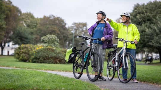 Two smiling, chatting women stand in a tree-lined park holding their bicycles and looking off to the left.