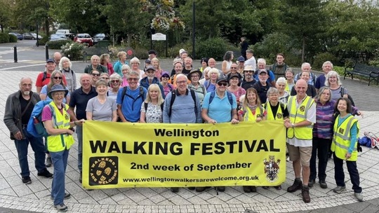 A crowd of people in outdoor clothing standing outside Wellington Leisure Centre with a banner that reads Wellington Walking Festival