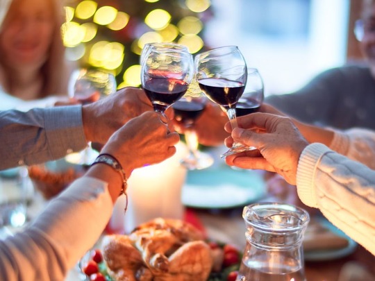 A table spread with a festive meal, over which people are clinking their glasses together. There's a Christmas tree behind them.