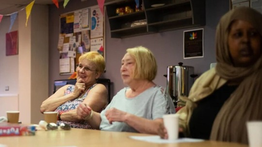 3 women sitting at a table talking with others out of view. The table has board games and drinks cups on it.