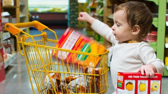 A toddler putting a box into a yellow toy shopping trolley in a supermarket.