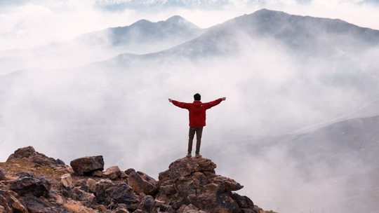 A person in a red coat, standing at the top of a rocky outcrop in the mountains, arms spread wide.