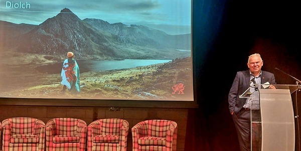 Rob standing at lectern on stage infront of screen with image of mountains and word Diolch