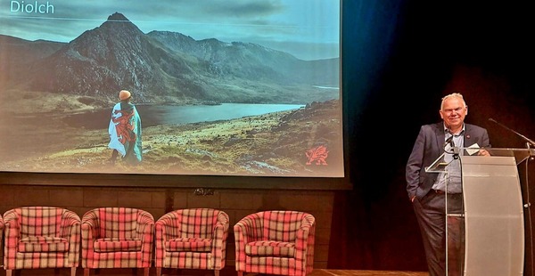 Rob standing at lectern on stage infront of screen with image of mountains and word Diolch
