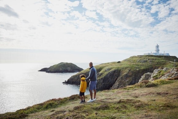 Dyn a bachgen yn sefyll ar lwybr arfordir Cymru, yn syllu allan ar arfordir Gogledd Cymru, a’r môr eang yn ymestyn i’r gorwel.