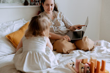 Parent and child on a bed; the parent is pointing at a laptop as they speak to the child